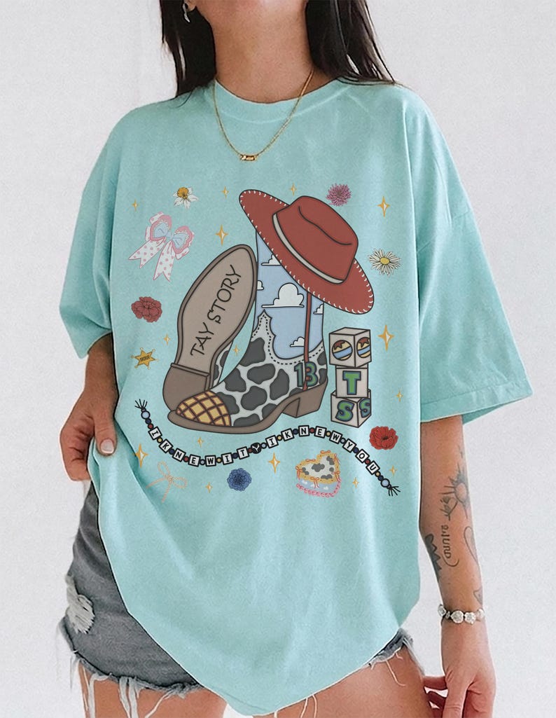 May include: A light blue t-shirt featuring a graphic print of a cowboy boot with a cow print pattern, a red cowboy hat, building blocks with the letters T, S, and the number 13, and a beaded necklace graphic with the text I KNEW IT I KNEW YOU.