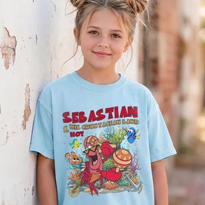 以下が含まれることがあります： 水色のTシャツで、マラカスを演奏するカニのセバスチャンと「Sebastian & His Crustacean Band Hot」という文字が描かれたカラフルなグラフィックプリントが特徴です。周囲にはサンゴ、魚、錨が鮮やかな漫画風のスタイルで描かれています。
