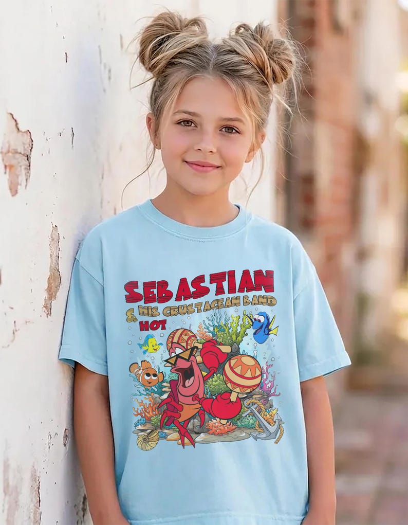 以下が含まれることがあります： 水色のTシャツで、マラカスを演奏するカニのセバスチャンと「Sebastian & His Crustacean Band Hot」という文字が描かれたカラフルなグラフィックプリントが特徴です。周囲にはサンゴ、魚、錨が鮮やかな漫画風のスタイルで描かれています。
