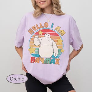 May include: Orchid-colored t-shirt featuring a graphic of Baymax from Big Hero 6. The design includes the text "HELLO I AM BAYMAX" in a colorful, retro style. The shirt has short sleeves and a relaxed fit.