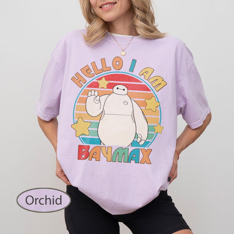 May include: Orchid-colored t-shirt featuring a graphic of Baymax from Big Hero 6. The design includes the text "HELLO I AM BAYMAX" in a colorful, retro style. The shirt has short sleeves and a relaxed fit.