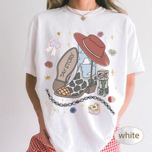 May include: A white short-sleeve t-shirt featuring a graphic print of a cowboy boot with cow print, a red hat, alphabet blocks, a beaded bracelet with the text I KNEW IT, and the text TAY STORY. The design includes small flowers and stars.