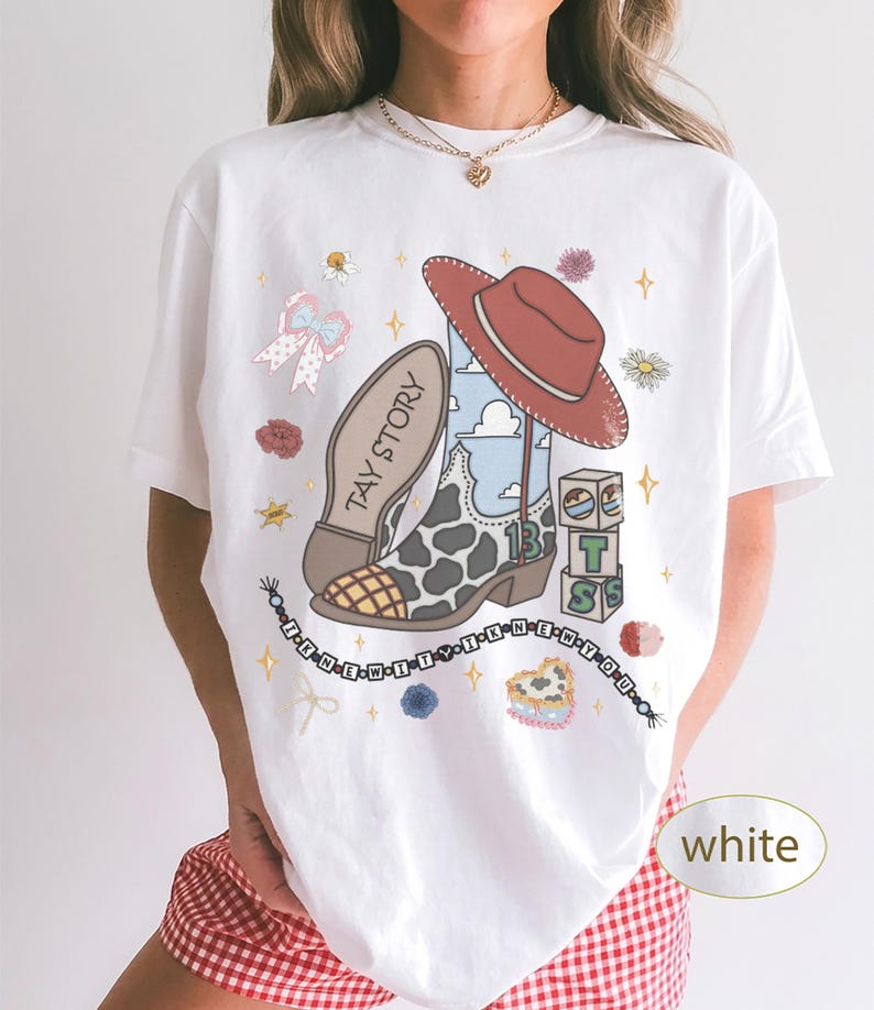 May include: A white short-sleeve t-shirt featuring a graphic print of a cowboy boot with cow print, a red hat, alphabet blocks, a beaded bracelet with the text I KNEW IT, and the text TAY STORY. The design includes small flowers and stars.