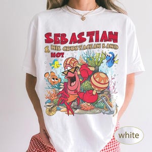 以下が含まれることがあります： マラカスを演奏するセバスチャンのキャラクターと「SEBASTIAN & HIS CRUSTACEAN BAND HOT」というテキストがプリントされた白いTシャツ。デザインには、色鮮やかな海中のサンゴ、カクレクマノミ、ナンヨウハギ、錨が描かれています。