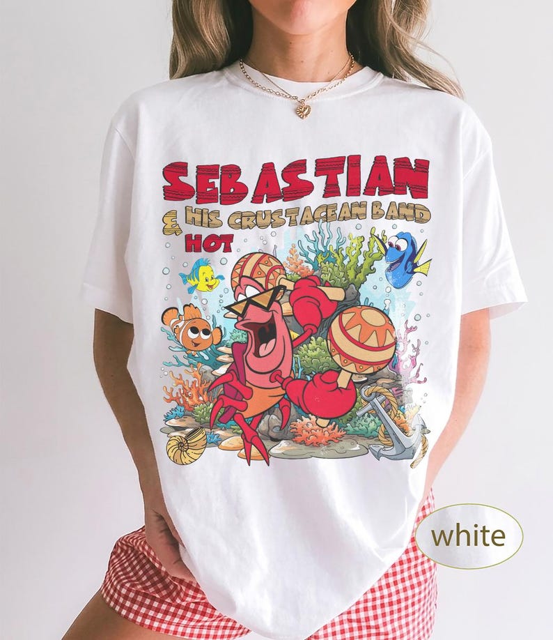 以下が含まれることがあります： マラカスを演奏するセバスチャンのキャラクターと「SEBASTIAN & HIS CRUSTACEAN BAND HOT」というテキストがプリントされた白いTシャツ。デザインには、色鮮やかな海中のサンゴ、カクレクマノミ、ナンヨウハギ、錨が描かれています。