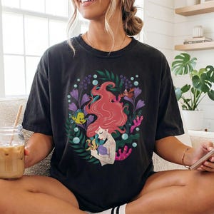 Peut inclure: T-shirt noir avec une illustration color&eacute;e d'une sir&egrave;ne aux cheveux roux, entour&eacute;e de plantes aquatiques et de cr&eacute;atures marines. Le motif comprend un poisson jaune, des fleurs violettes et du corail. Le t-shirt est de coupe ample.