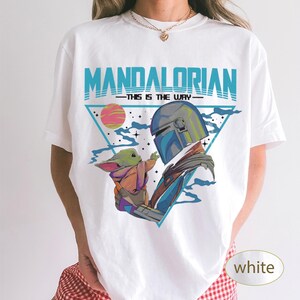 Pu&ograve; includere: Una t-shirt bianca con una stampa grafica di The Mandalorian e Grogu. Il design include il testo "MANDALORIAN" e "THIS IS THE WAY" in blu, su uno sfondo triangolare con un pianeta rosa, stelle e nuvole blu.
