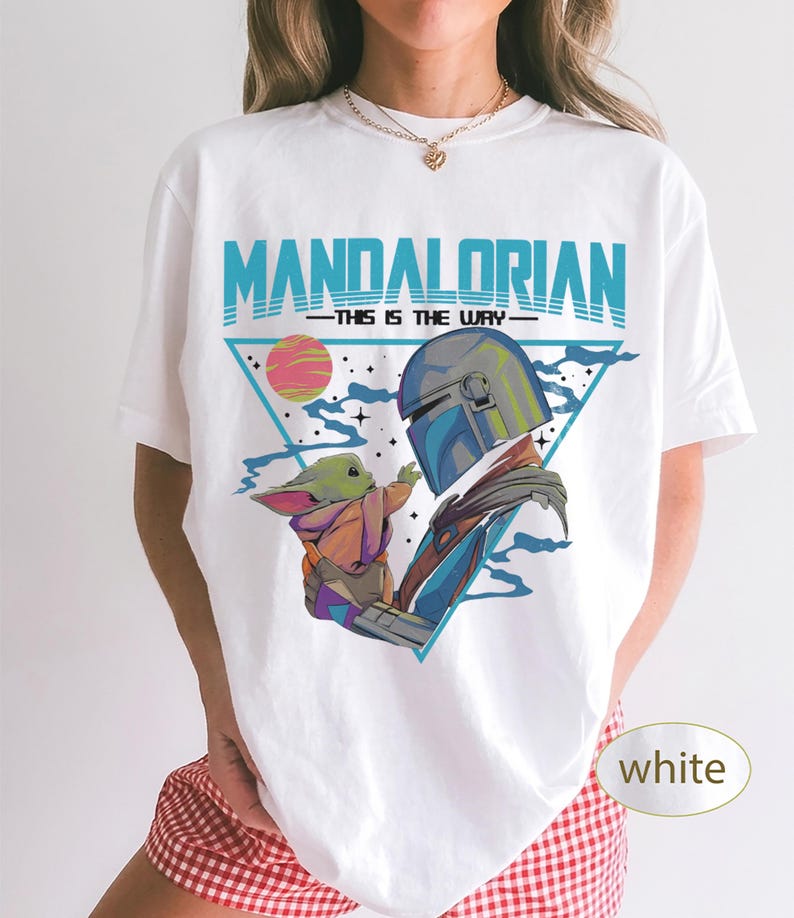 Pu&ograve; includere: Una t-shirt bianca con una stampa grafica di The Mandalorian e Grogu. Il design include il testo "MANDALORIAN" e "THIS IS THE WAY" in blu, su uno sfondo triangolare con un pianeta rosa, stelle e nuvole blu.