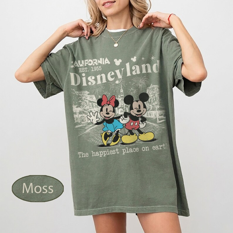 May include: Moss green t-shirt featuring a graphic of Mickey and Minnie Mouse with the text "California Disneyland Est. 1955" and "The happiest place on earth." The shirt has a vintage, distressed look.