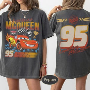 May include: Dark gray t-shirt featuring a graphic of Lightning McQueen from the movie Cars. The front design includes the number 95, checkered flags, and the text "Lightning McQueen". The back of the shirt also displays "95" and "Lightning McQueen".
