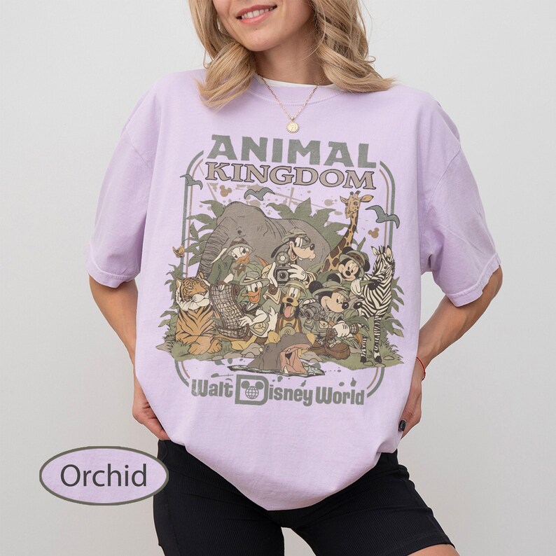 May include: Orchid-colored t-shirt featuring a graphic of various cartoon animals, including an elephant, giraffe, and zebra, with the text "Animal Kingdom" and "Walt Disney World". The shirt has short sleeves and a crew neck.