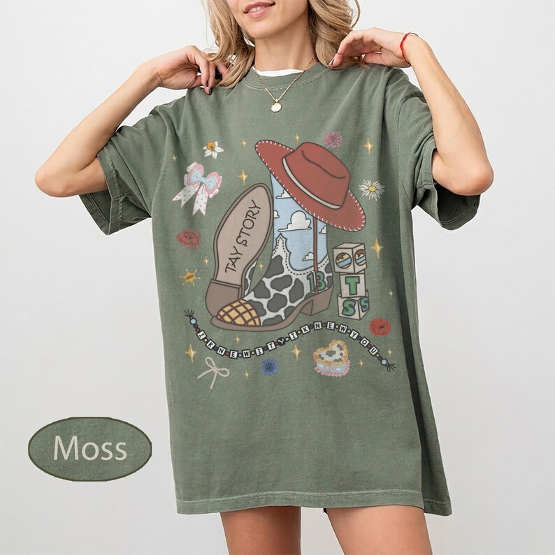 May include: A moss green short-sleeve t-shirt featuring a graphic print of a cowboy boot with a cow print pattern, a brown hat, and stacked blocks with the letters T, S. The text TAY STORY is printed on the boot, surrounded by small floral icons.