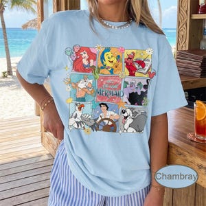 May include: Pale blue t-shirt with a colourful graphic of characters from "The Little Mermaid." The design includes Ariel, Sebastian, Flounder, and other characters in a grid pattern. The shirt has short sleeves and a relaxed fit.