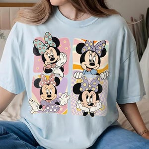 May include: Light blue t-shirt featuring four panels with Minnie Mouse illustrations. Each panel showcases a different pose of Minnie Mouse with polka dot bows and outfits, set against pastel backgrounds.