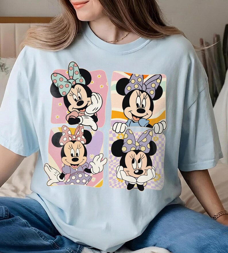 May include: Light blue t-shirt featuring four panels with Minnie Mouse illustrations. Each panel showcases a different pose of Minnie Mouse with polka dot bows and outfits, set against pastel backgrounds.