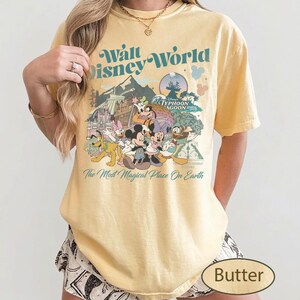 May include: Butter-colored t-shirt featuring a graphic of Disney World characters and the text "Walt Disney World" and "The Most Magical Place On Earth." The design includes Mickey Mouse, Minnie Mouse, Donald Duck, and other characters.