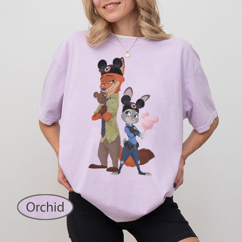 Pu&ograve; includere: T-shirt color orchidea con un'illustrazione a cartoni animati di Nick Wilde e Judy Hopps di Zootropolis. Nick indossa una camicia verde e cravatta, Judy un'uniforme da poliziotta. Entrambi i personaggi indossano cappelli con orecchie da topo.