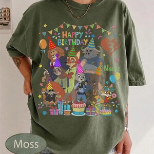May include: Moss green t-shirt featuring a colorful graphic of Zootopia characters celebrating a birthday. The design includes the text "Happy Birthday" and a number 3. Balloons, presents, and a cupcake are also visible.