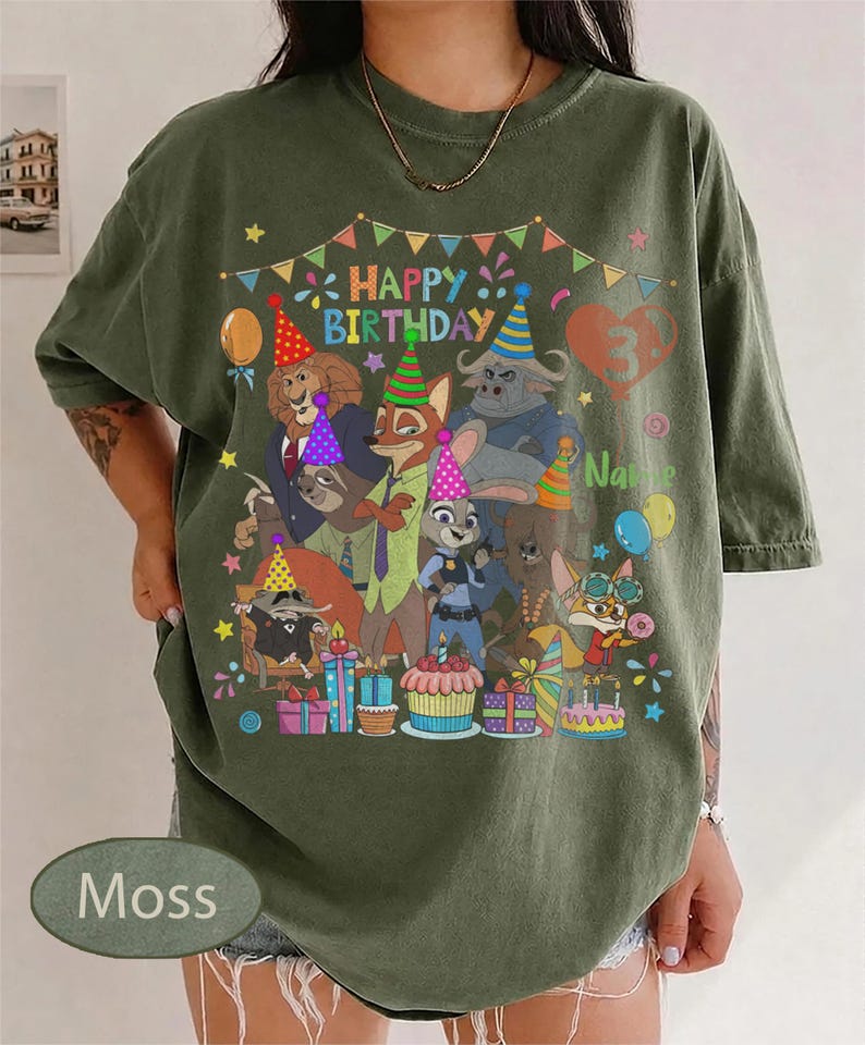 May include: Moss green t-shirt featuring a colorful graphic of Zootopia characters celebrating a birthday. The design includes the text "Happy Birthday" and a number 3. Balloons, presents, and a cupcake are also visible.