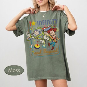 May include: Moss green t-shirt featuring a graphic of Buzz Lightyear and Woody from Toy Story, with the text "Infinity and Beyond". The design includes a star, rocket, and other colorful elements. The shirt has short sleeves and a relaxed fit.