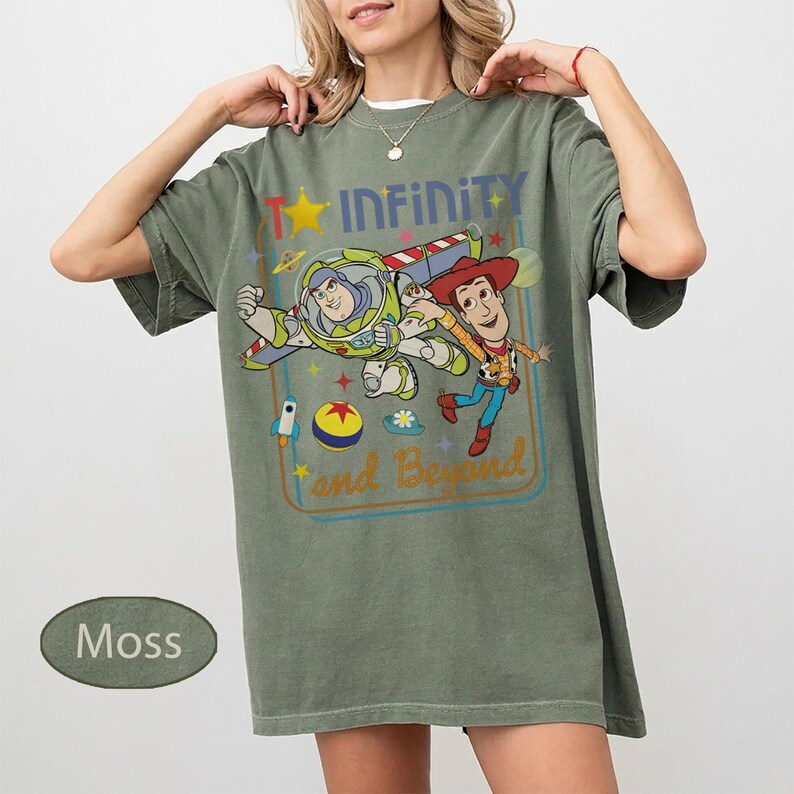 May include: Moss green t-shirt featuring a graphic of Buzz Lightyear and Woody from Toy Story, with the text "Infinity and Beyond". The design includes a star, rocket, and other colorful elements. The shirt has short sleeves and a relaxed fit.