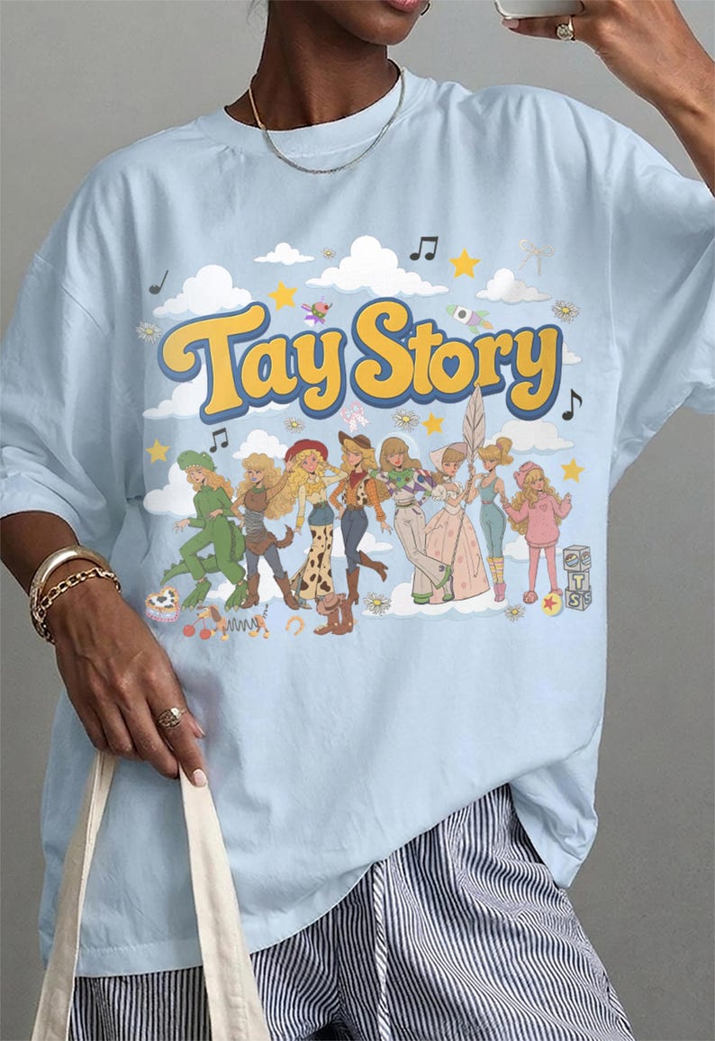 K&ouml;nnte beinhalten: Ein hellblaues T-Shirt mit dem Schriftzug Tay Story in gelben Buchstaben mit blauem Umriss. Unter dem Text befinden sich acht illustrierte Figuren in verschiedenen Outfits, umgeben von Wolken, Sternen, Musiknoten und kleinen Spielzeugsymbolen.