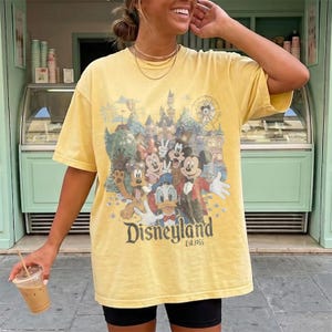 May include: A pale yellow short-sleeve t-shirt featuring a vintage-style graphic print of Mickey Mouse, Minnie Mouse, Donald Duck, Goofy, and Pluto in front of a castle. The text Disneyland Est. 1955 is printed in a dark font below the characters.