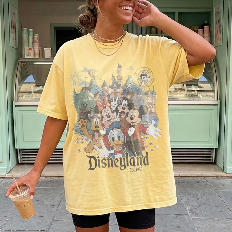 May include: A pale yellow short-sleeve t-shirt featuring a vintage-style graphic print of Mickey Mouse, Minnie Mouse, Donald Duck, Goofy, and Pluto in front of a castle. The text Disneyland Est. 1955 is printed in a dark font below the characters.