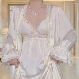 Puede incluir: Conjunto de lencería color crema con un vestido lencero de satén, un bralette con ribete de encaje y una bata a juego. La bata tiene mangas con volantes y detalles de encaje. Un collar de perlas añade un toque de elegancia al conjunto.