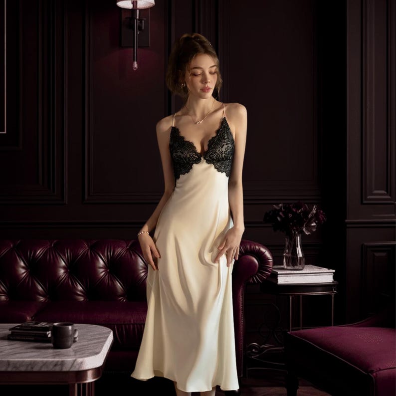 Puede incluir: Vestido lencero de sat&eacute;n color crema con detalles de encaje negro en el corpi&ntilde;o. El vestido tiene tirantes finos y largo midi. La imagen est&aacute; ambientada en una habitaci&oacute;n con paneles de madera oscura y un sof&aacute; burdeos.