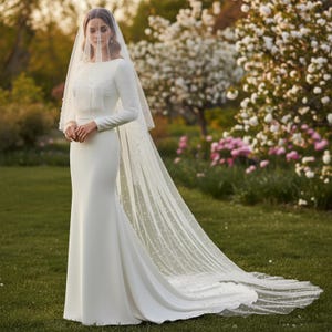 May include: A white wedding dress with long sleeves and a floor-length skirt. The dress features a simple, elegant design with a fitted bodice and a flowing skirt. A sheer veil, adorned with small, scattered pearls, drapes over the dress and trails behind.