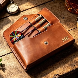 May include: A tan leather dart case with a snap closure and the embossed letters MK, containing three darts with metallic barrels and colourful, multi-patterned flights. The case is constructed with individual slots for each dart.