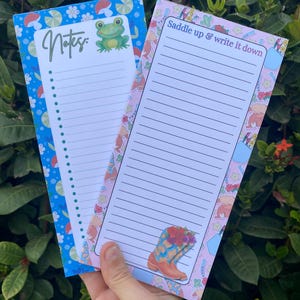 May include: Two rectangular notepad sheets with lined paper. The left sheet has a blue border with mushrooms and flowers, the text "Notes:", and a frog illustration. The right sheet has a pink border with cowboy boots and the text "Saddle up & write it down".