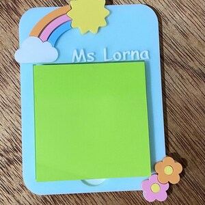 May include: A light blue memo pad with a green sticky note, a rainbow, a yellow sun, and two flowers. The text "Ms Lorna" is written in white. The memo pad is on a wooden surface.