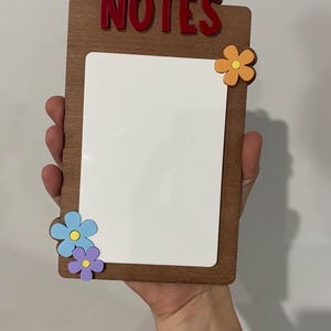 May include: A wooden memo board with the word "NOTES" in red letters at the top. The board has a white writing surface and is decorated with colourful flower accents. The board is being held by a person.