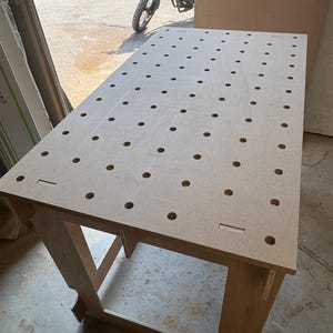 May include: A light brown workbench with numerous evenly spaced holes across the surface. The workbench has a rectangular shape and is supported by four legs. The legs are connected by a cross brace. A motorcycle is visible in the background.