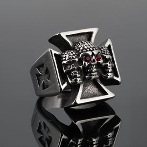 Op de afbeelding: Zilverkleurige ring met een Maltezer kruisontwerp en drie schedels met rode edelsteenogen. De ring heeft een gepolijste afwerking en een gedetailleerd ontwerp. Een opvallend sieraad.