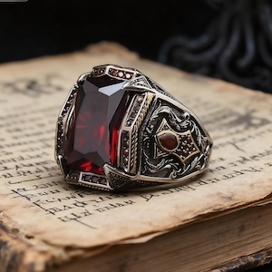 May include: A silver ring with a large, rectangular, deep red gemstone as the centerpiece. The ring has intricate detailing with smaller red stones and gold accents. The ring is set on a vintage paper background.