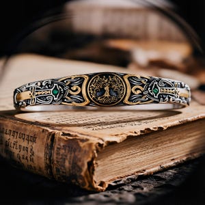 Puede incluir: Una pulsera plateada y dorada con un intrincado diseño de nudo celta. La pulsera presenta un emblema central del Árbol de la Vida y pequeñas piedras preciosas verdes. La pulsera está sobre un libro antiguo abierto.