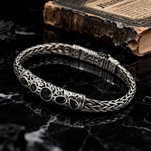 May include: A silver bracelet featuring a woven chain design and a central decorative panel. The panel displays intricate scrollwork engravings and is set with five smooth, black, teardrop and circular stones arranged in a linear pattern.