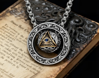 May include: Silver-tone necklace featuring a pendant with an all-seeing eye design. The pendant has a gold-tone triangle with a blue gem, set within a decorative circular frame. The necklace rests on an open book with aged pages.