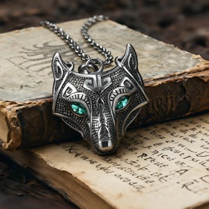 Puede incluir: Un collar con colgante de lobo plateado con un diseño detallado. Los ojos del lobo son de color verde esmeralda. El colgante cuelga de una cadena plateada. El collar descansa sobre un libro abierto y envejecido con texto descolorido.