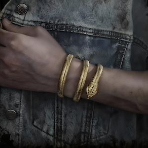 May include: A gold snake bracelet coiled around a wrist. The bracelet has a detailed snake head and a textured body. The jewellery is worn over a denim jacket.