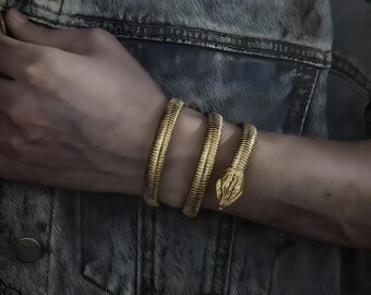 May include: A gold-colored snake bracelet coiled around a wrist. The bracelet features a detailed snake head design and a textured body. The jewelry is worn over a denim jacket.