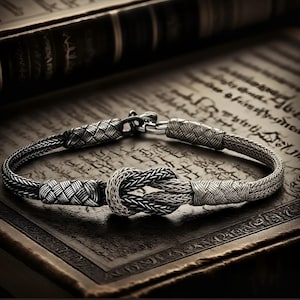 Pode incluir: Uma pulseira de prata com um design entrançado e um elemento central em forma de nó. A pulseira é feita de um metal brilhante e é exibida em uma superfície escura e texturizada, possivelmente um livro. Fecho de encaixe.