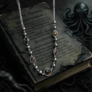May include: A silver necklace with oval, amber-colored stones and small silver beads. The necklace rests on an open, aged book with gothic script. A dark octopus sculpture is in the background.