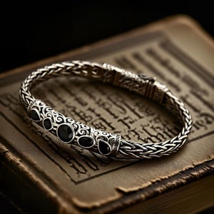 Puede incluir: Una pulsera de plata con un diseño de cadena tejida y detalles de piedras preciosas negras. La pulsera presenta detalles intrincados y se muestra en un libro antiguo.