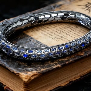May include: A silver bracelet with a hexagonal pattern and blue gemstones. The bracelet is resting on an open antique book with aged pages and dark leather cover. The bracelet's design is reminiscent of a snake.