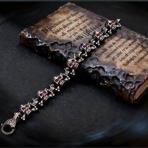 May include: A silver bracelet featuring a series of skull charms with red accents. The bracelet is displayed on a dark surface next to a vintage book with aged pages and faded text. The clasp has a decorative design.