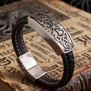 May include: A black leather bracelet with a silver rectangular centrepiece. The centrepiece features Celtic knot designs and a compass-like symbol. The bracelet has a braided leather band and a magnetic clasp.